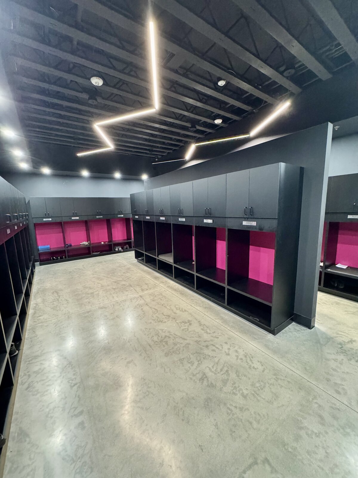 Rows of black athlete lockers with hot pink back panels, name tags, bench seating and shoe shelves under zigzagging LED ceiling lights