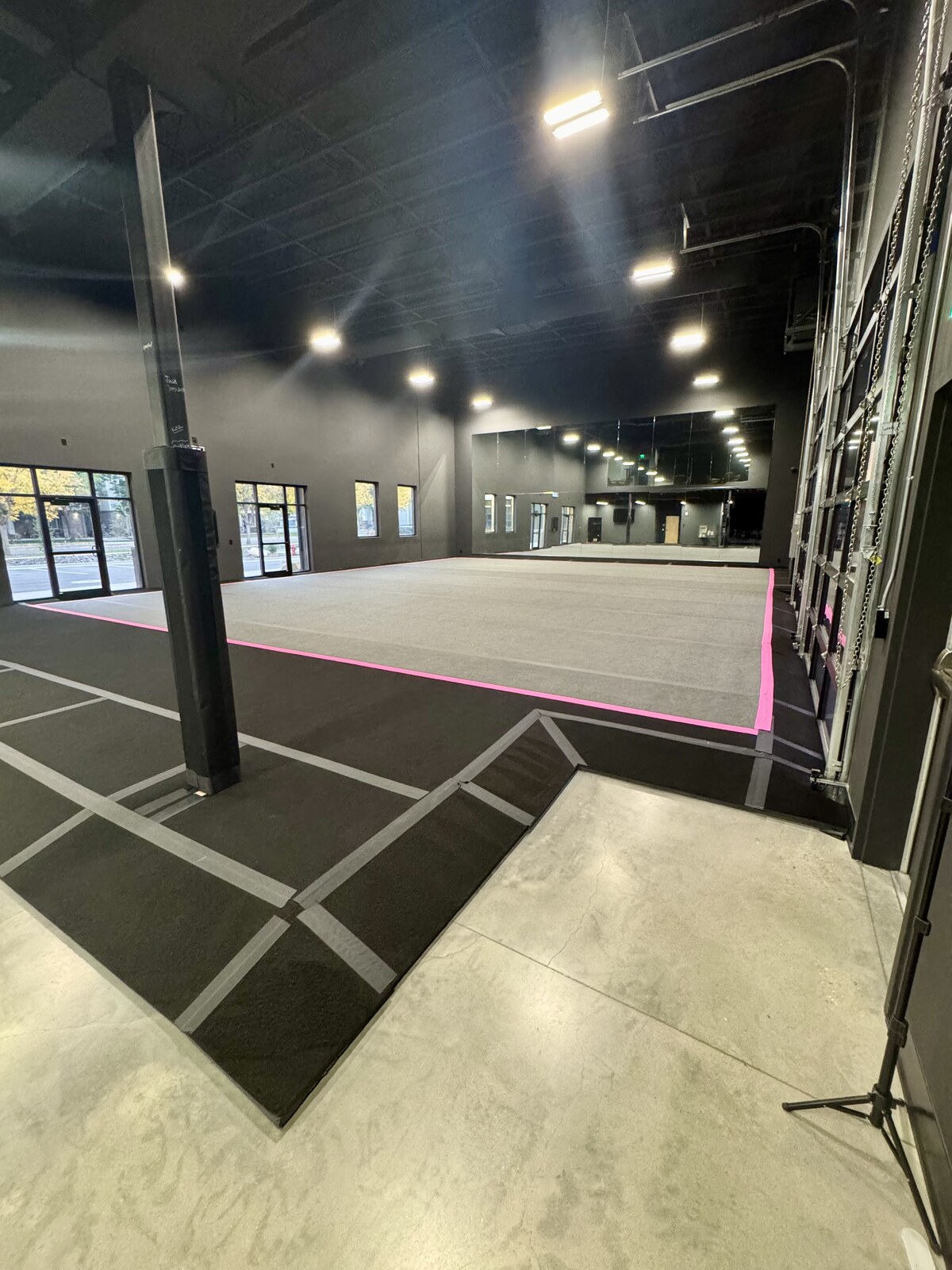 Another full cheer floor, with a gray performance mat edged in hot pink tape, a mirrored back wall, windows along the left, and a glass roll-up door on the right