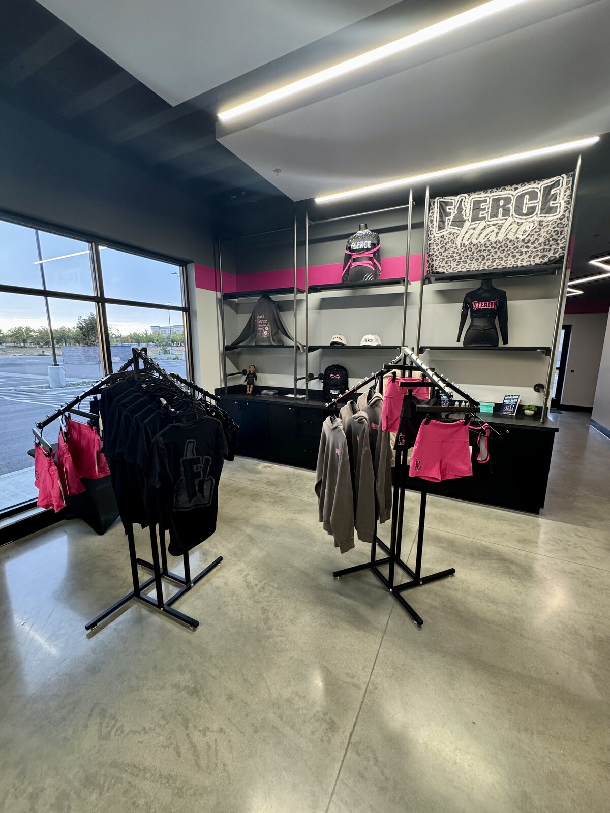 The Fierce Pro Shop, with garment racks of black and pink apparel in front of matte black shelving carrying Fierce Idaho banners, mannequins, caps, and bags beneath a hot pink accent stripe