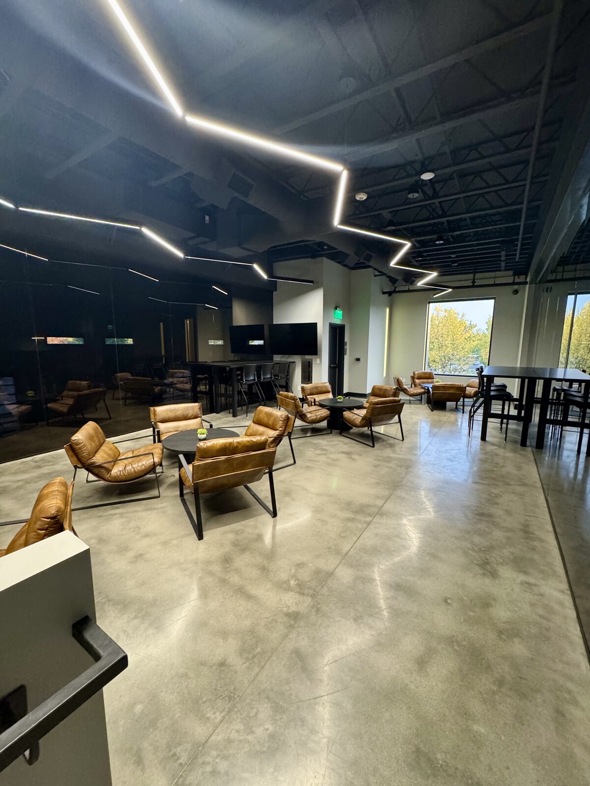 The parent viewing lounge from another angle, showing clusters of tan leather armchairs, tall dark bar tables with stools, and a large window looking out onto sunlit trees